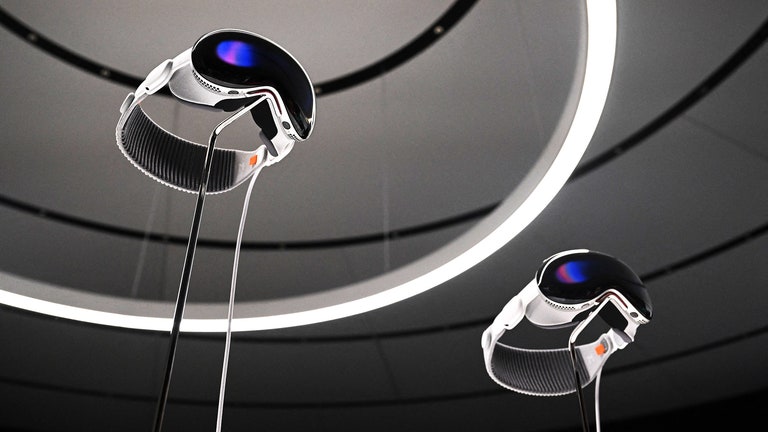 Two Apple Vision Pro headsets mounted on display stands shot from below with circular geometric shapes on the ceiling...